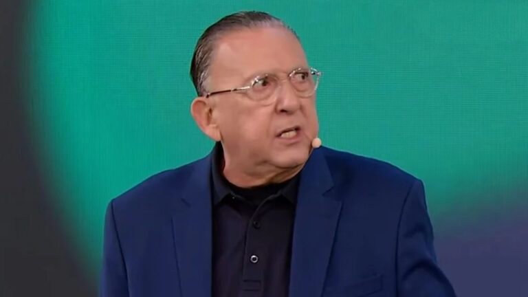 Homem de óculos e cabelo penteado para trás fala com expressão séria, usando paletó azul-escuro e camisa polo preta. Ele está com um microfone de lapela preso ao rosto, diante de um fundo verde e azul em estúdio de televisão.