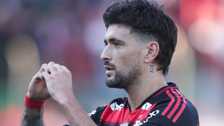 Jogador Giorgian de Arrascaeta de perfil com as mãos juntas fazendo sinal de coração veste camisa do Flamengo em campo de futebol