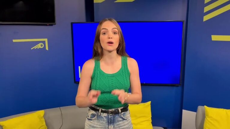 Mulher com blusa verde fala gesticulando em estúdio com sofá cinza e tela azul ao fundo