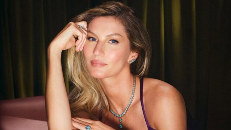 Mulher posando de forma elegante, com expressão serena e olhar direto para a câmera. Ela usa joias sofisticadas — colar, brincos e anéis — e está vestida com uma roupa de alça fina em tom escuro. O fundo é composto por cortinas em tons de verde e dourado, criando uma atmosfera luxuosa e intimista.