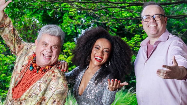 Três pessoas sorridentes posam ao ar livre, vestindo roupas coloridas e brilhantes, com árvores verdes ao fundo