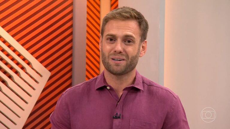 Homem com barba curta e cabelos claros veste uma camisa social roxa e fala em frente a um fundo com listras diagonais laranjas e pretas, além de elementos geométricos brancos, com o logotipo da TV Globo visível no canto inferior direito da imagem.