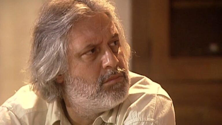 Homem de cabelos grisalhos e barba espessa aparece em close com expressão séria, olhando para o lado. Ele veste camisa bege clara e está em ambiente interno com iluminação suave e tons amarelados.