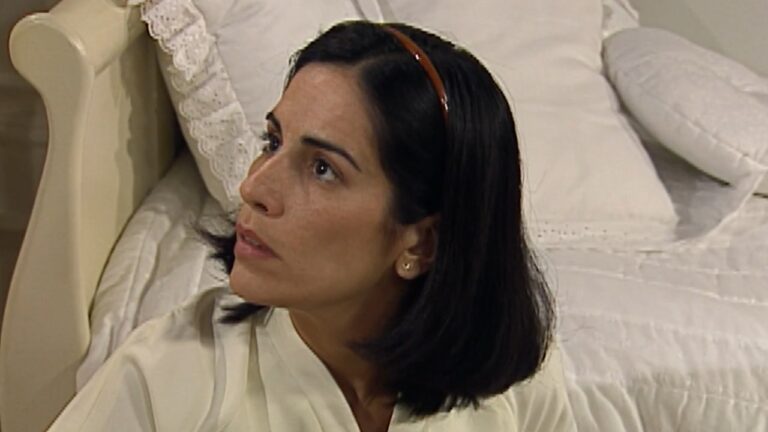 Mulher de cabelos escuros na altura dos ombros usa tiara fina e roupa clara enquanto está sentada no chão ao lado de uma cama com colcha branca e detalhes rendados olhando para cima com expressão apreensiva em quarto iluminado por luz suave com móveis claros e ambiente doméstico organizado