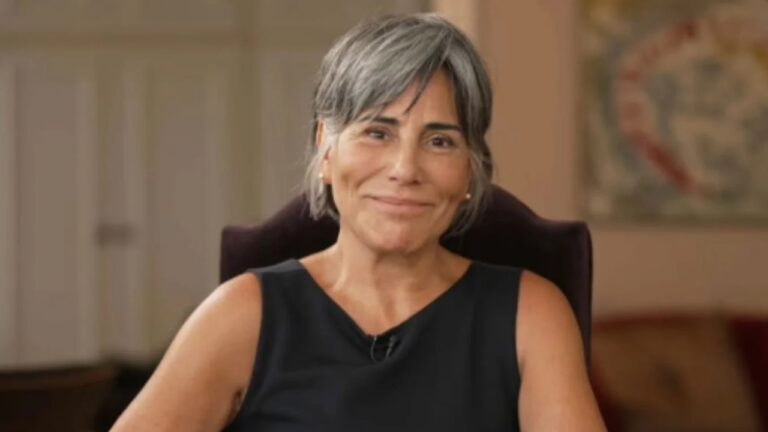 Mulher de cabelos curtos grisalhos e lisos sorri levemente enquanto está sentada em uma cadeira de encosto alto. Ela usa um vestido preto sem mangas e brincos discretos. O ambiente ao fundo é interno, com tons suaves e um quadro desfocado na parede.