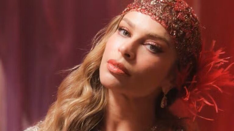 Mulher posa com expressão elegante, usando tiara ornamentada com pedrarias e penas vermelhas, maquiagem marcante e cabelo loiro ondulado, em cenário iluminado por luz avermelhada.