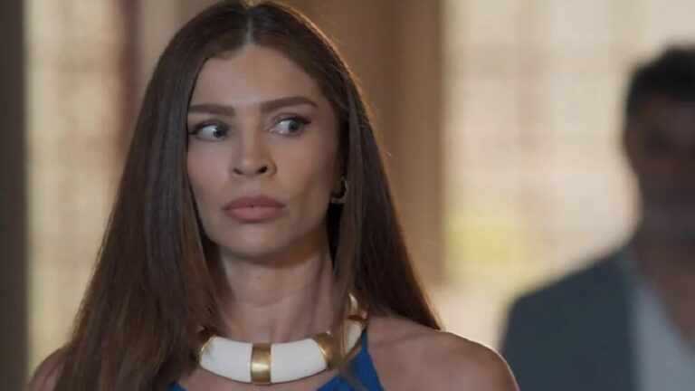 Mulher com expressão de surpresa ou desconfiança, olhando para o lado. Ela tem cabelos lisos e compridos, usa um vestido azul e um colar branco com detalhes dourados. Ao fundo, há uma figura masculina desfocada e um ambiente interno bem iluminado.