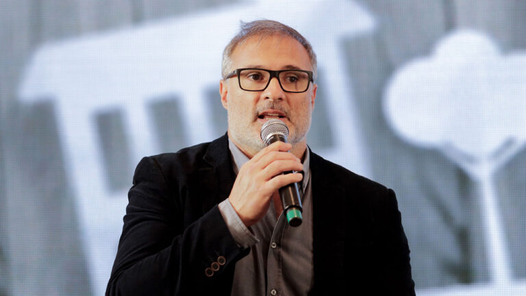 Guillermo Pendino fala ao microfone com óculos de armação preta e paletó escuro durante evento com telão digital ao fundo