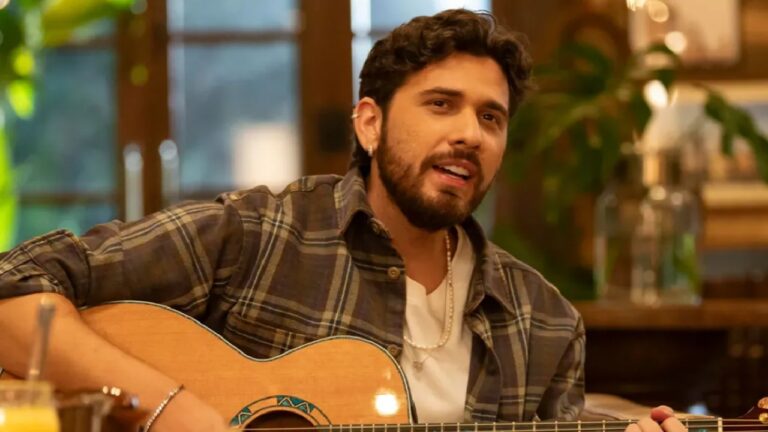 Homem toca violão e canta em ambiente interno iluminado com luz amarelada. Ele usa camisa xadrez sobre camiseta branca, colar de pérolas e brinco pequeno, com expressão concentrada enquanto segura o instrumento. Ao fundo, há plantas, móveis de madeira e janelas grandes com vista para o exterior.