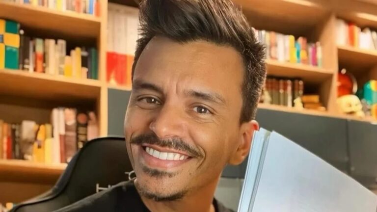 Homem sorri segurando um livro aberto em ambiente com estante repleta de livros ao fundo, iluminado por luz quente.