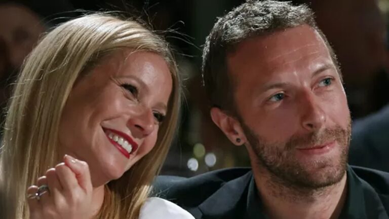 Mulher e homem estão sentados lado a lado, sorrindo e aparentando cumplicidade. Ela tem cabelos loiros lisos, usa batom vermelho e apoia o queixo na mão, inclinando-se levemente em direção a ele. O homem tem barba curta, cabelo castanho e veste camisa escura, olhando para frente com expressão serena. O fundo está desfocado, sugerindo um ambiente de evento ou jantar.