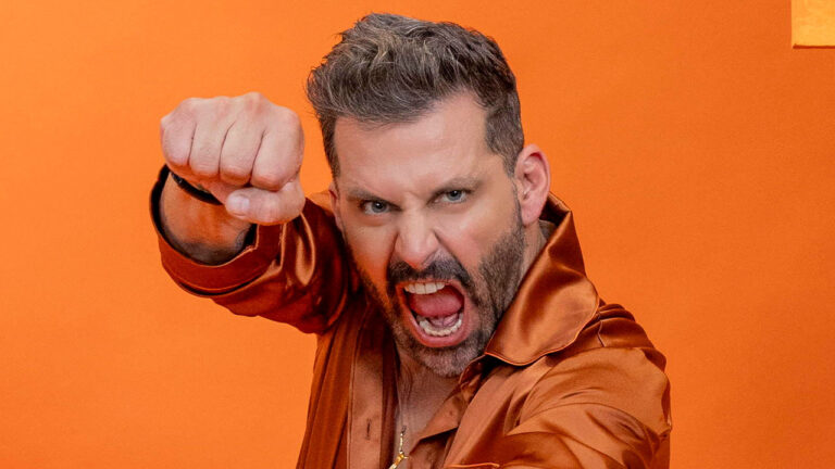 Henri Castelli encara a câmera com expressão de fúria e punho cerrado para frente em ensaio fotográfico sobre fundo laranja