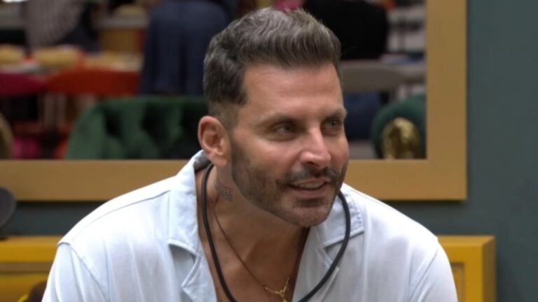 Homem de barba e cabelos curtos sorri enquanto conversa, usando camisa clara e colar, em um ambiente interno colorido que lembra uma casa de reality show.