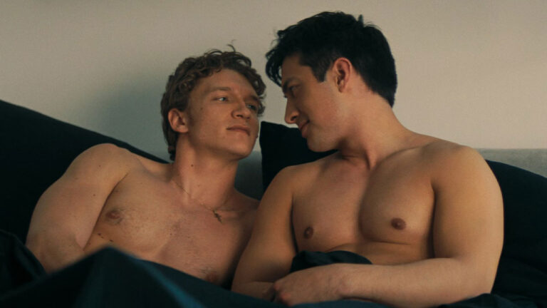 Ilya Rozanov (Connor Storrie) e Shane Hollander (Hudson Williams) sem camisa trocam olhares carinhosos na cama na série Heated Rivalry