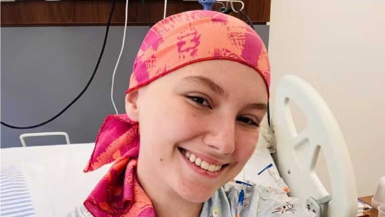 Mulher jovem sorri tirando uma selfie em um quarto de hospital. Ela usa um lenço colorido em tons de rosa e laranja na cabeça e veste uma roupa hospitalar. Atrás dela há uma cama branca, equipa
