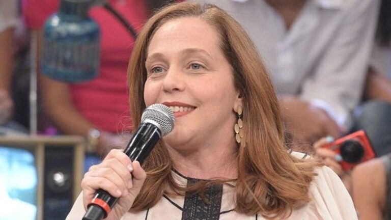 Mulher de cabelos castanhos médios segura um microfone e sorri enquanto fala, usando blusa clara e brincos dourados, em ambiente de estúdio com pessoas desfocadas ao fundo assistindo.