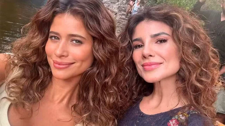 Duas mulheres com cabelos cacheados castanhos sorriem para uma selfie ao ar livre em local com vegetação e rio ao fundo. A mulher à esquerda usa blusa clara e a da direita veste roupa escura com bordados florais coloridos. Ambas aparecem iluminadas pela luz natural, com expressão alegre.