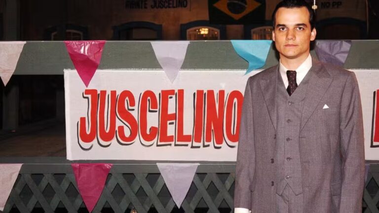 Homem de terno cinza posa ao lado de faixa com o nome Juscelino em cenário decorado com bandeirinhas coloridas