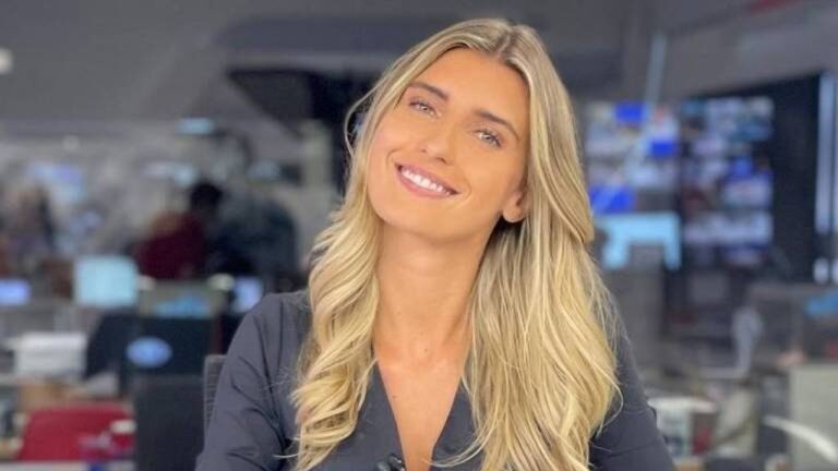 Mulher de cabelos loiros compridos e ondulados sorri levemente enquanto está sentada em um ambiente de redação jornalística com telas e pessoas desfocadas ao fundo. Ela veste uma blusa escura e olha diretamente para a câmera em um cenário iluminado e profissional.