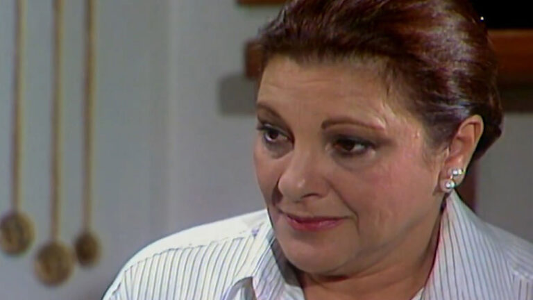 Yara Amaral como Joana na novela Fera Radical usa camisa branca listrada e brincos de pérola com expressão pensativa em cena da trama