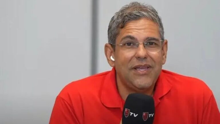 Homem com cabelos grisalhos curtos e óculos fala segurando um microfone preto com o logotipo da FlaTV. Ele usa camisa polo vermelha e fones de ouvido brancos, em frente a um fundo neutro cinza claro.