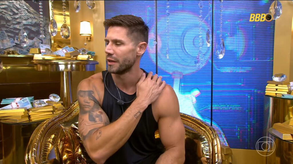 Jonas Sulzbach, com expressão séria está sentado em uma poltrona dourada dentro de um ambiente luxuoso, decorado com barras douradas, cédulas de dinheiro e cristais pendurados. Ele veste regata preta e toca o ombro, enquanto fala no confessionário do programa BBB 26