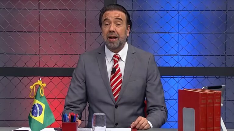 Jorge Lordello, vestindo um terno cinza e gravata vermelha com listras brancas, está sentado em uma mesa do cenário do Operação de Risco, da RedeTV!. À sua frente há uma pequena bandeira do Brasil, canetas e livros vermelhos. O fundo é composto por uma parede com padrão de grade metálica iluminada por luzes nas cores azul e vermelha, criando um visual contrastante