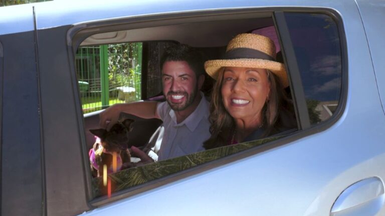Homem e mulher sorriem dentro de um carro, vistos pela janela aberta; a mulher usa chapéu de palha e o homem segura um pequeno cachorro de colo com roupa colorida.