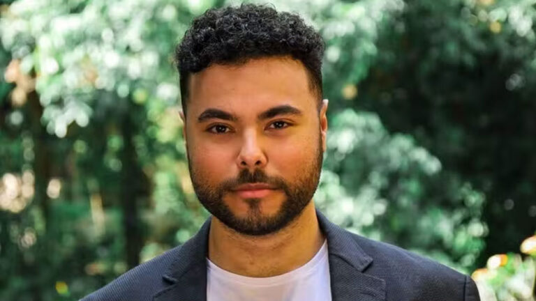 Juan Jullian encara a câmera com expressão séria usando blazer escuro sobre camiseta branca com fundo de folhagens verdes desfocadas