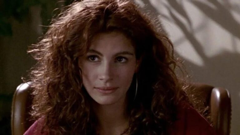 Mulher com cabelos cacheados castanho-avermelhados e expressão serena está sentada em cadeira de couro, levemente inclinada para frente. Ela usa brincos de argola e roupa em tom escuro, olhando com leve sorriso. O fundo mostra parede clara com sombras projetadas de plantas.