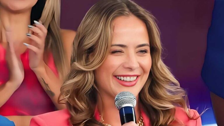 Mulher de cabelos ondulados sorri ao segurar microfone em palco enquanto público aplaude ao fundo em evento com iluminação colorida