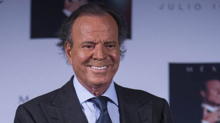Julio Iglesias sorri em evento público usando terno escuro e gravata diante de painel com fotos e nomes ao fundo