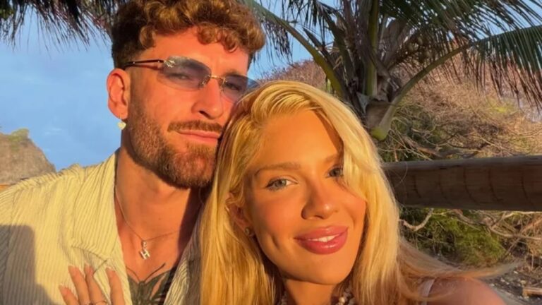 Casal posa sorridente em selfie ao pôr do sol, com vegetação e coqueiros ao fundo; o homem usa óculos escuros e camisa clara, e a mulher tem cabelo loiro solto
