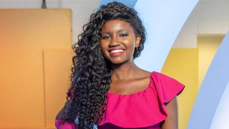 Mulher sorridente posa para a câmera usando um vestido rosa de um ombro só, em frente a um fundo colorido com tons de azul e amarelo, em ambiente interno bem iluminado.