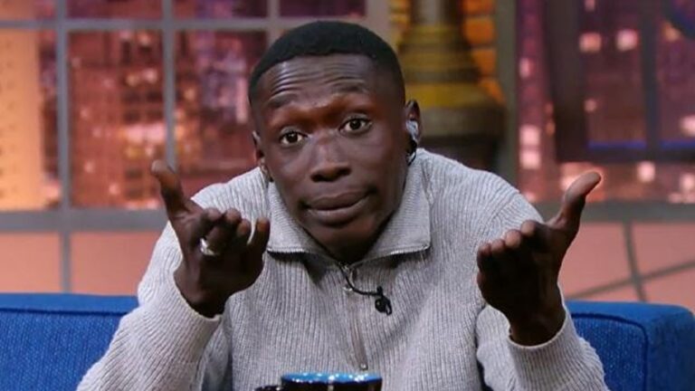 Homem sentado em programa de entrevistas faz gesto com as mãos abertas e expressão divertida, usando suéter cinza claro e fone de ouvido