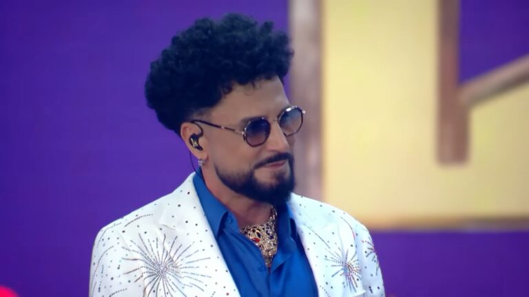 A imagem mostra o cantor Latino, que é um homem com barba e cabelo cacheado, usando óculos escuros e brinco, durante uma apresentação musical no SBT. Ele veste uma camisa azul e um paletó branco decorado com brilhos e desenhos de fogos de artifício, além de um colar dourado. O fundo é roxo e iluminado, de um evento musical