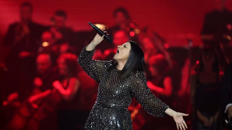 Mulher canta com intensidade em um palco iluminado por luz vermelha, segurando um microfone com uma das mãos enquanto estende a outra, vestindo um vestido preto brilhante com mangas longas, diante de uma orquestra desfocada ao fundo.