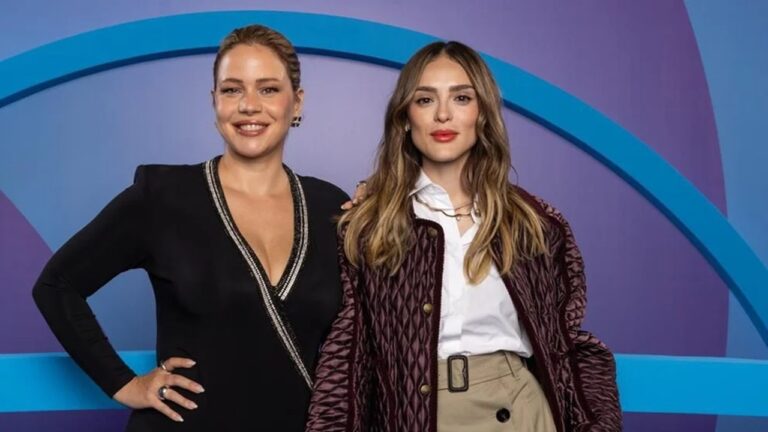 Duas mulheres posando lado a lado diante de um fundo roxo e azul com formas geométricas. A mulher à esquerda veste um vestido preto com decote em V e detalhes brilhantes, sorrindo e com uma das mãos na cintura. A mulher à direita usa uma camisa branca, casaco vinho acolchoado e calça bege, olhando para a câmera com expressão confiante e batom vermelho.