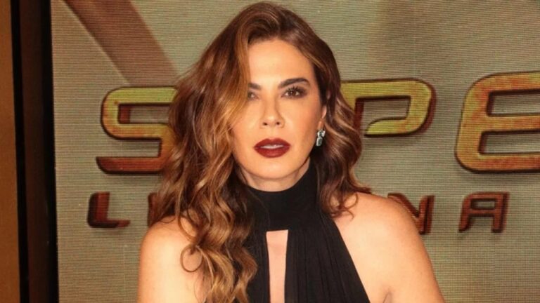 Mulher de cabelos ondulados castanhos usa vestido preto sem mangas diante de painel luminoso com logotipo de evento