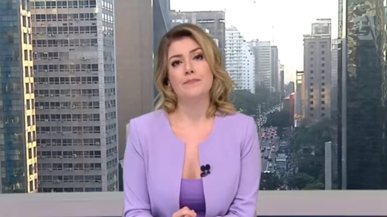 Mulher apresentadora de telejornal com expressão séria, vestindo blazer lilás e blusa roxa, usando microfone de lapela preso à roupa. Ao fundo, há uma grande janela mostrando prédios altos e uma avenida movimentada com carros em fila, sugerindo uma grande cidade.