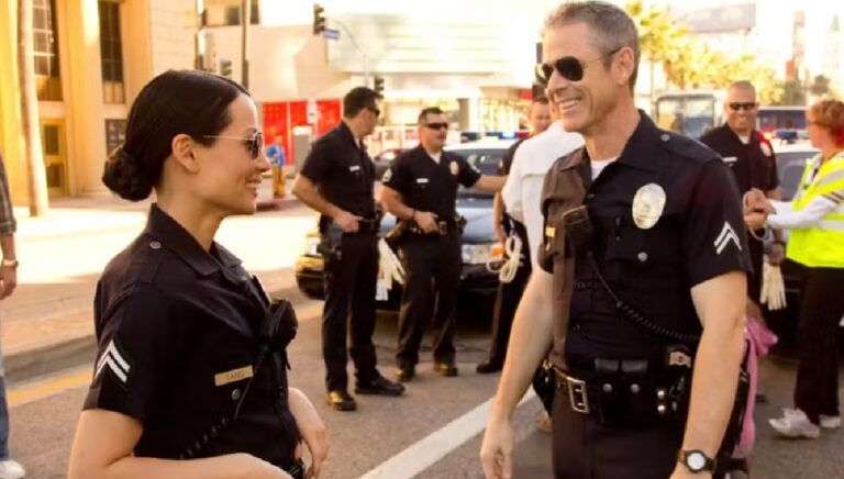 Homem e mulher vestindo uniformes de polícia conversam sorrindo em uma rua durante o dia. Ambos usam óculos escuros e rádios presos ao uniforme. Ao fundo, outros policiais estão reunidos próximos a viaturas com luzes ligadas, e algumas pessoas observam a cena.