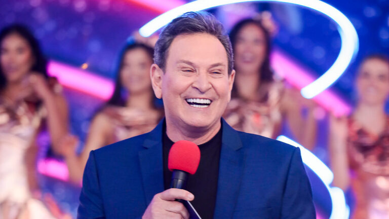 Apresentador Luís Ricardo sorri com blazer azul e segura microfone vermelho no palco do SBT com bailarinas ao fundo do estúdio