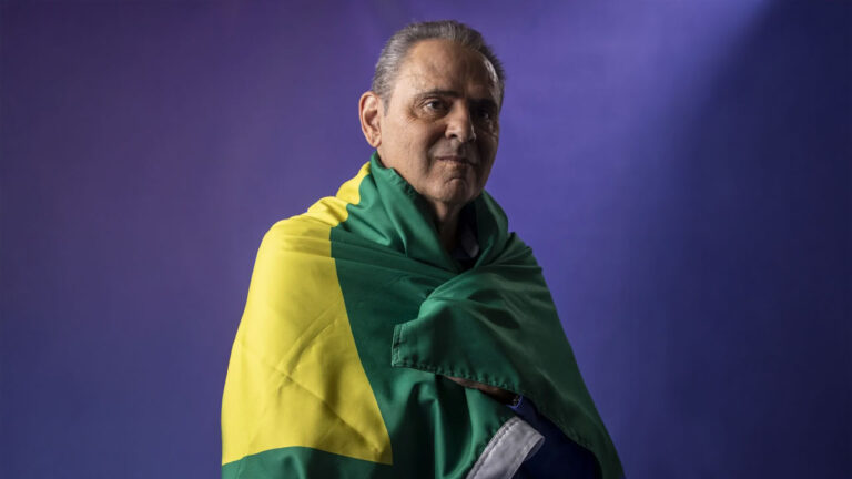 O narrador Luis Roberto posa com a bandeira do Brasil enrolada sobre os ombros em frente a um fundo roxo para cobertura esportiva na Globo