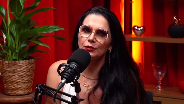 Luiza Ambiel de óculos e batom vermelho fala ao microfone de podcast em estúdio com fundo vermelho e planta ornamental ao lado
