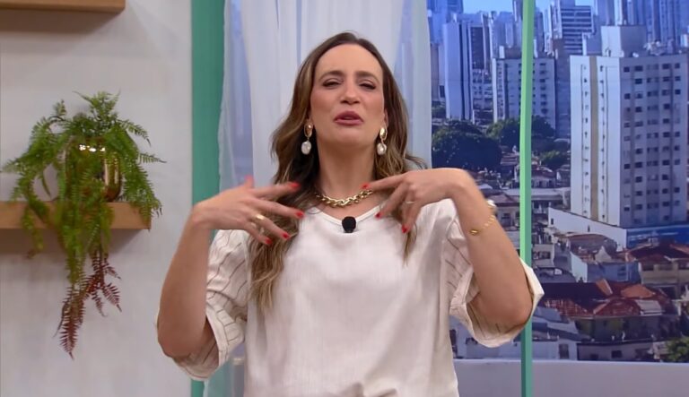 Mulher em estúdio de televisão gesticula com as mãos erguidas e expressão animada enquanto fala. Ela usa blusa clara de mangas médias, colar dourado grosso e brincos com pérolas pendentes. Ao fundo, há uma janela grande com vista para prédios de uma cidade e uma parede com prateleira contendo vaso dourado com planta verde.