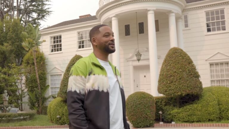 Homem de barba curta e cabelo raspado nas laterais sorri enquanto está em frente a uma grande mansão branca de estilo clássico, com colunas altas e janelas simétricas. Ele veste uma jaqueta verde e preta com camiseta branca, e ao redor há jardins bem cuidados com arbustos podados e árvores decorativas.