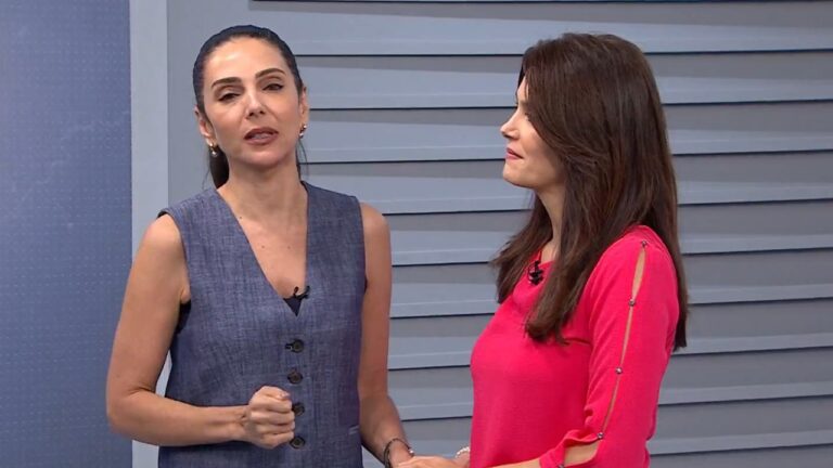 Duas mulheres adultas em estúdio de televisão em plano médio a mulher à esquerda tem pele clara cabelos escuros presos para trás maquiagem discreta expressão séria e fala olhando levemente para a câmera vestindo colete sem mangas em tom cinza com botões frontais e microfone preso à roupa com a mão direita parcialmente fechada em gesto de ênfase a mulher à direita tem pele clara cabelos castanhos longos soltos expressão atenta olhando para a mulher ao lado vestindo blusa rosa de mangas