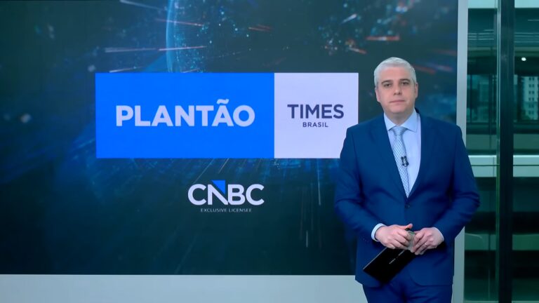 Marcelo Favalli, vestindo um terno azul-claro e gravata, aparece em estúdio de telejornal em frente a um telão com os logotipos Plantão Times Brasil e CNBC Exclusive Licensee. O cenário é moderno e corporativo, com iluminação fria e vista parcial de janelas ao fundo