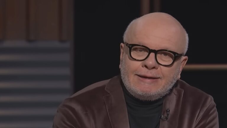 Homem careca com barba grisalha curta e óculos de armação grossa escura aparece em ambiente interno de iluminação suave. Ele veste suéter preto de gola alta e blazer marrom, olhando ligeiramente para a câmera com expressão tranquila. O fundo mostra parede com listras horizontais escuras.