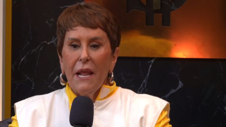 Mulher fala ao microfone em ambiente interno com fundo escuro e iluminação alaranjada, vestindo roupa branca e amarela com detalhes dourados e brincos grandes.